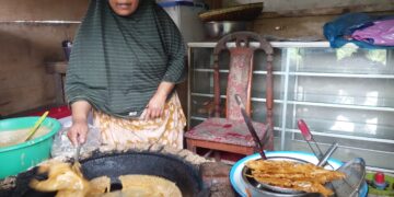 Seorang pedagang gorengan sedang memasak dengan menggunakan wajan besar di dapur sederhana